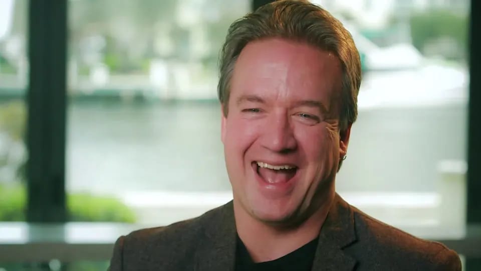 Testimonial video still of an interviewee laughing, with a waterfront view behind him